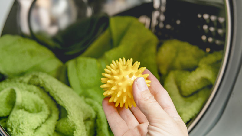 Uma mão de adulto introduz numa máquina de secar roupa uma bola de plástico amarela onde já se encontra uma toalha de turco verde.
